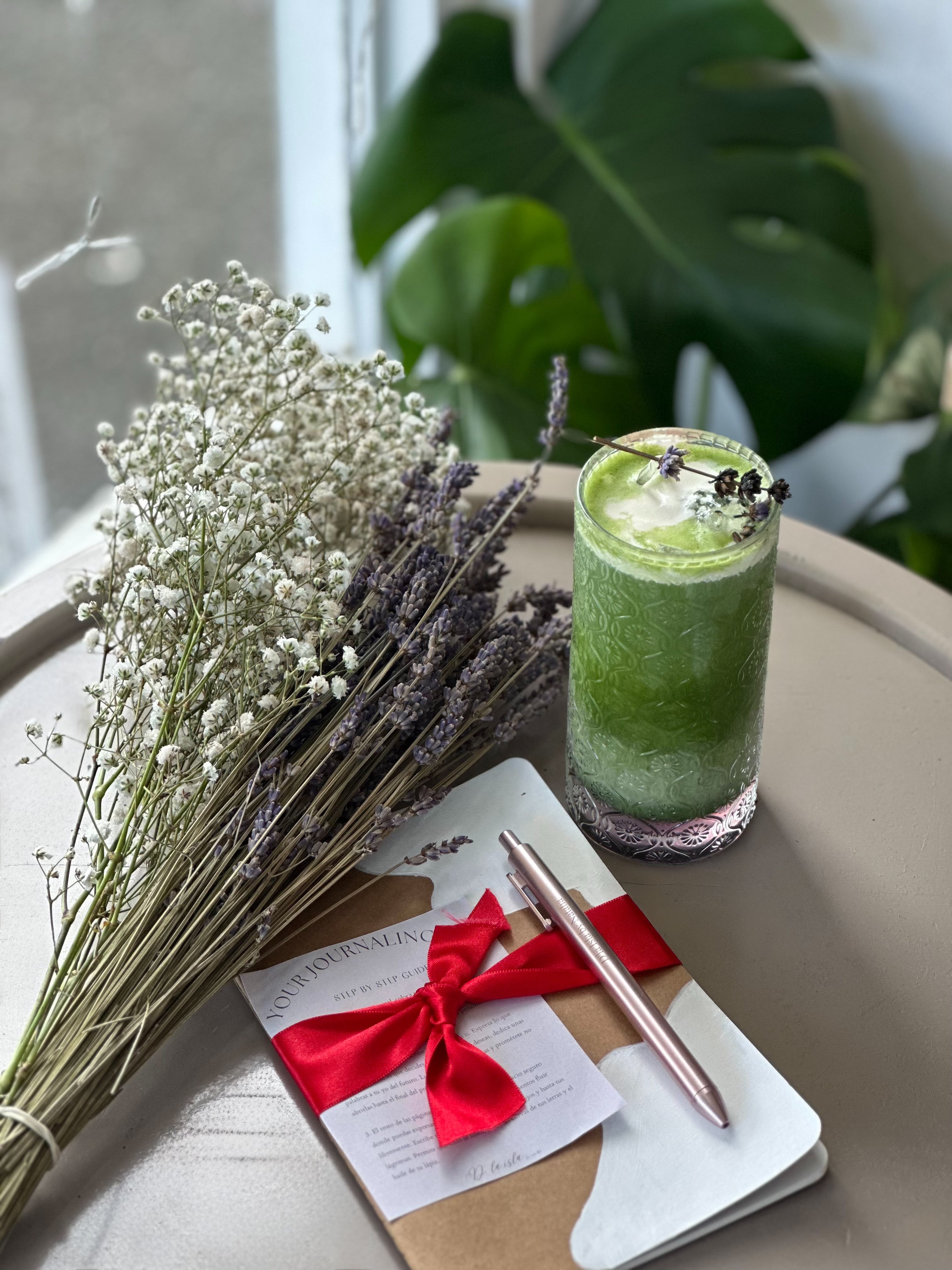 A styled flat-lay featuring a green iced drink in a clear glass, a bouquet of dried white baby's breath and lavender, a kraft paper notepad tied with a red ribbon, and a rose gold pen on a neutral tabletop with a green plant leaf in the background.