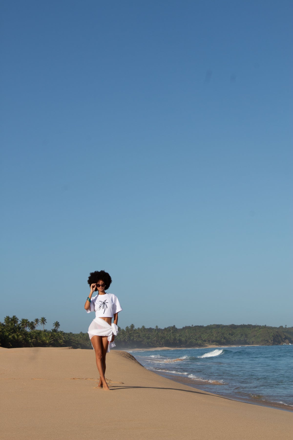 A person stands on a sandy beach facing the ocean under a clear blue sky, wearing a white short-sleeve crop top and a white skirt.